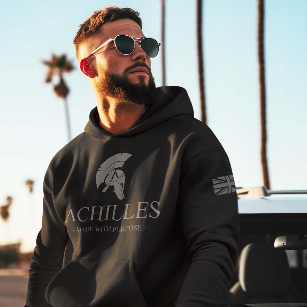 Front view of a man sat on a car wearing a unsiex fit black cotton pull over hoodie with grey Achilles logo screen printed across the front